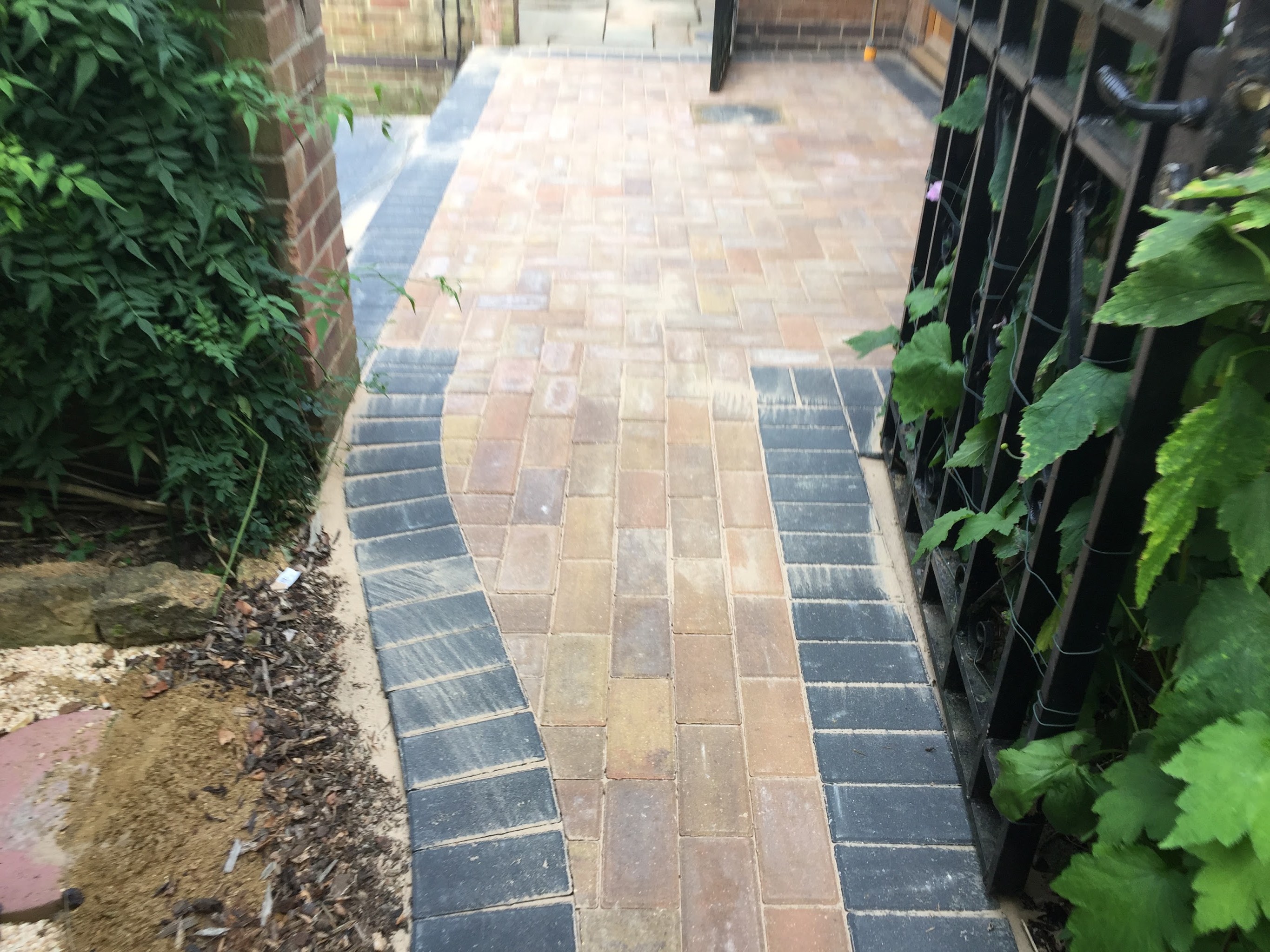 Block-paved side path with curved charcoal borders beside an iron railing and vine